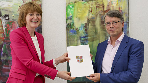 Bowen_Hennemeyer Die beiden Personen stehen vor einer Wand mit zwei großformatigen Bildern. Sie halten gemeinsam die Ernennungsurkunde in der Hand.