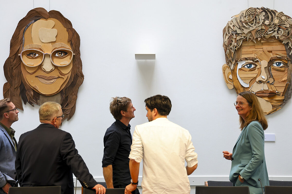 KMK-Ausstellung An einer Wand hängen zwei überlebensgroße Porträts einer weiblichenund einerr männlichen PErsonaus verschiedenfarbigen Holzteilen. Davor stehen die genannten Personen und unterhalten sich.