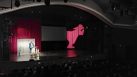 Es ist eine Theaterbühne zu sehen. Dort steht ein Mann an einem Pult. Links hinter ihm befinden sich Banner mit kleinen Fischer-Logos und dem Schriftzug "Jahresempfang 2024". Im rechten Bildteil ist ein fast fünf Meter hohes Fischerlogo auf dem roten Theatervorhang zu sehen.  