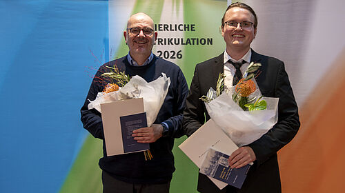 Zwei Männer in Anzügen stehen nebeneinander, halten jeweils einen Blumenstrauß und eine Urkunde, im Hintergrund ein Banner mit dem Text 'FEIERLICHE MATRIKULATION 2026'