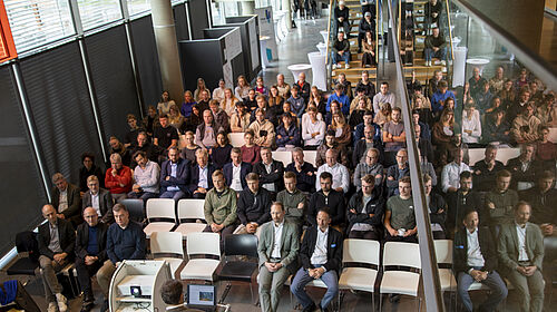 Blick von oben auf ein Auditorium mit zahlreichen sitzenden Personen bei einer Tagung, vorne steht ein Redner an einem Pult mit Laptop und Beamer.