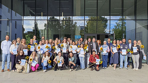 Eine Gruppe von Menschen steht oder hockt in mehreren Reihen vor einem modernen Glasgebäude. Viele halten Urkunden und Sonnenblumen in den Händen.